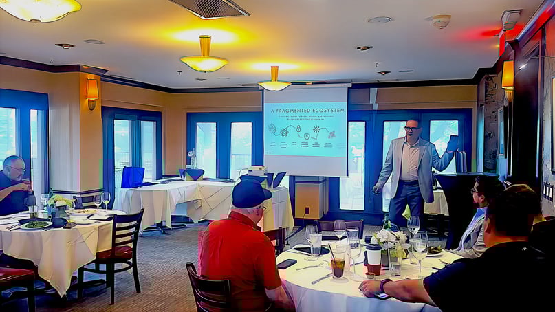 Business professionals seated at round tables during a wireless technology lunch meeting, with a speaker presenting in front of a screen titled "A Fragmented Ecosystem."